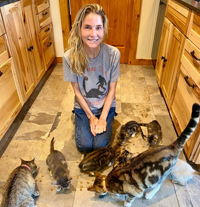 Elizabeth Wing Carlisle kneeling on a kitchen floor, smiling, with a group of cats and kittens eating from the tiles around her